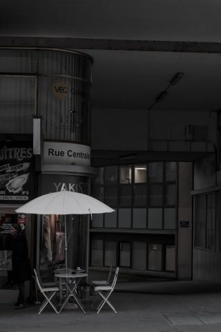 Petite terrasse de rue avec un parasol blanc et des chaises sur un trottoir urbain sombre à l'angle de la Rue Centrale.