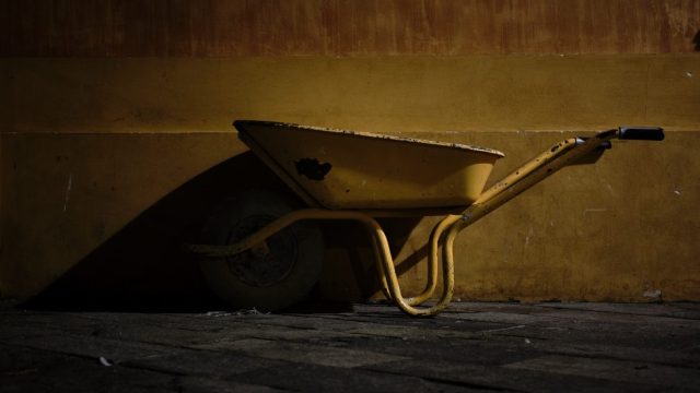 Brouette de chantier jaune stationnée contre un mur aux tons ocre, créant un fort contraste avec des ombres dramatiques.