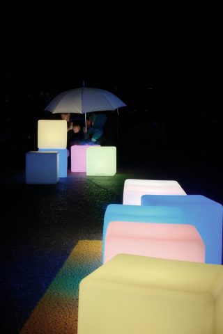Installation artistique de cubes lumineux colorés éclairant la rue la nuit, avec une personne assise sous un parapluie