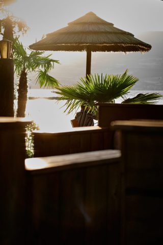 Coucher de soleil paisible sur un lac vu depuis une terrasse estivale, avec un parasol en paille et des palmiers en contre-jour