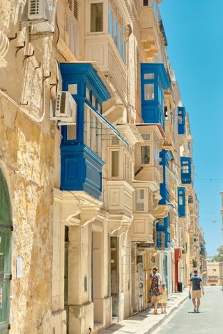 Ruelle typique de La Valette à Malte avec ses balcons en bois peints en bleu et blanc et des passants qui marchent au soleil.