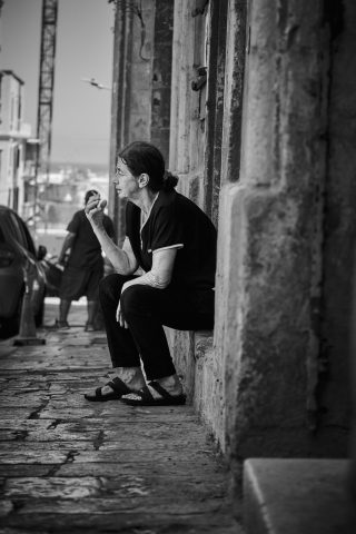 Photo de rue en noir et blanc d'une habitante assise sur le pas d'une porte en pierre et fumant une cigarette à Malte.