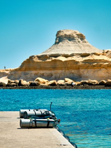 Bouteilles de plongée sous-marine alignées sur un quai en béton face à une eau turquoise et au rocher de Fungus Rock à Gozo, Malte.