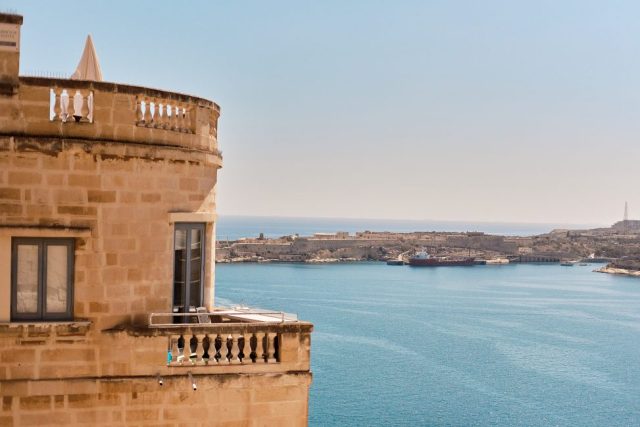 Façade en pierre maltaise avec un balcon donnant sur une baie d'eau bleue et des fortifications historiques à Malte.